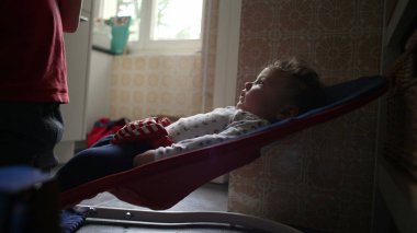 Bebek bir fedaide yatar, ağabeyi yanında dururken sakince yukarı bakar, rahat bir ev ortamı, mutfakta nazik bir kardeş etkileşimi.