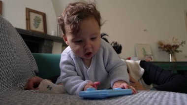 Bebek, mavi nesneyle oynarken koltukta öne eğiliyor, odaklanmış ifade, merakla bebek keşif nesnesi, kapalı alanda eğlenceli bir an, yakınlarda annesiyle aile bağı kurma zamanı.