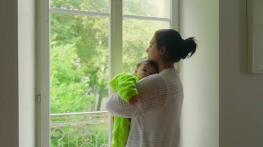 Anne pencerenin yanında duruyor, bebek omzunda dinleniyor, anne derin düşüncelere dalıyor, bebek yavaşça uykuya dalıyor, sakin bir atmosferde sakin bir bağ kuruyor.