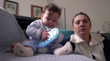 Bebek koltukta oturup mavi nesneyi çiğniyor, anne gözlemliyor, evde huzurlu bir aile anı yaşıyor, bebek oynuyor ve keşfediyor, bebekle anne arasında bağ kuruyor, oturma odası kuruyor.