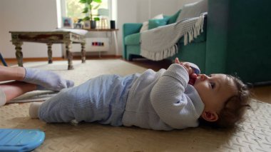 Bebek ağzında parmaklarıyla sırt üstü uzanıyor, sakin ve rahat bir yüz ifadesi, evde aile zamanı, bebek gezisi çevresi, huzurlu bir an, rahat bir evde neşeli bir çocuk.