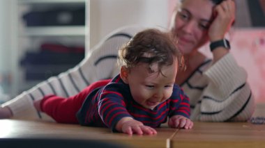 Bebek gülümsüyor ve masada emekliyor, neşeli ve enerjik, anne arka planda izliyor, bebek ve anne arasında canlı bir etkileşim, bebek anın tadını çıkarıyor.