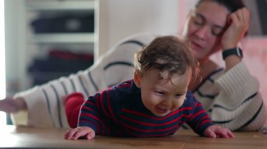 Bebek gülümsüyor ve masada emekliyor, neşeli ve enerjik, anne arka planda izliyor, bebek ve anne arasında canlı bir etkileşim, bebek anın tadını çıkarıyor.