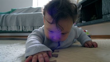 Bebek yerde yatıyor, yüzü yere yakın, tahta bir blokla etkileşimde, merakla çevreyi keşfediyor, ev ortamında güzel motor beceriler geliştiriyor.