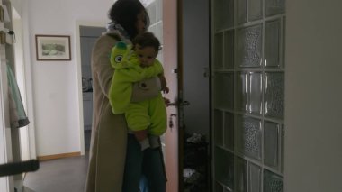 Anne bebeğini kucağında tutarken evden ayrılıyor, sıcak yeşil bir kıyafet giyiyor, dışarı çıkmaya hazırlanıyor, rahatlatıcı ve koruyucu bir jest yapıyor.