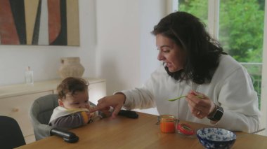 Anne bebeğini beslerken, sevgi dolu bir anı paylaşıyor, bebek gülümsüyor ve annesine bakıyor, yemek saatinde oyuncu etkileşimler, anne bebek mamasıyla kaşık tutuyor.