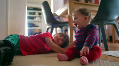 Bebek yerde oturuyor, ağabeyi yanında, bebek heyecanla gülümsüyor, evde oyun oynarken kardeş etkileşimi, kardeşle bebek arasında paylaşılan eğlenceli bir an.
