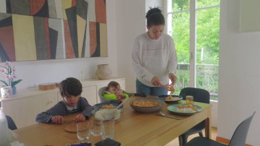 Anne yemek masasında yemek servisi yaparken, büyük çocuk ve bebek bakıcılığı, aile yemeği hazırlama ve sıcak bir evde toplanma.