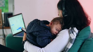 Anne kucağında uyuyan bir bebeği tutarken telefonunu kontrol eder ve dizüstü bilgisayarda çalışır, bakım ve uzaktan işleri harmanlar, ev ortamında birden fazla iş yapar.