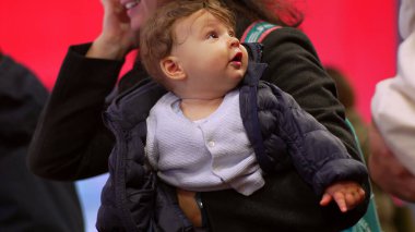 Bebek annesinin kollarında, yukarı bakıyor, kışlık ceket giyiyor, aile ortamını inceliyor, günlük sahne, ebeveyn-çocuk bağı, sıcak kıyafetler, kapalı bir spor salonu.