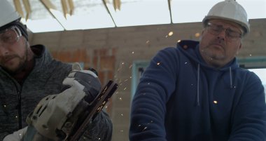 Koruyucu giysili iki adam, metal kesici ile metal kesiyor, kıvılcımlar inşaat alanını aydınlatıyor, takım çalışması ve hassasiyet süper yavaş çekimde.
