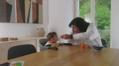 Anne bebeğini beslerken, sevgi dolu bir anı paylaşıyor, bebek gülümsüyor ve annesine bakıyor, yemek saatinde oyuncu etkileşimler, anne bebek mamasıyla kaşık tutuyor.