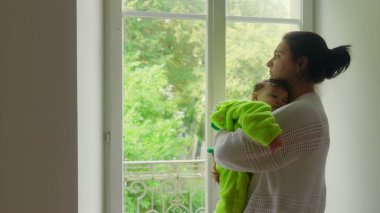 Anne pencerenin yanında duruyor, bebek omzunda dinleniyor, anne derin düşüncelere dalıyor, bebek yavaşça uykuya dalıyor, sakin bir atmosferde sakin bir bağ kuruyor.