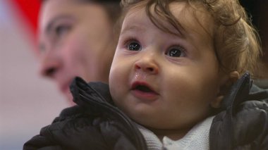 Bebek annesinin kollarında, açık gözlerle bakıyor, ceket giyiyor, tuhaf bir ifade takınıyor, günlük aile anı, yakın çekim görüntüsü, kapalı mekanda samimi etkileşim
