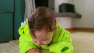 Bebek gülümsüyor ve yeşil bir kıyafetle yerde yatıyor, göbek zamanlarında çevreyle meşgul oluyor, neşeli ve eğlenceli, kapalı alanda oyun zamanı.