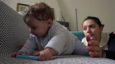 Bebek koltukta uzanmış mavi bir nesneyi tutarken, anne objeye odaklanıyor, oyun oynayarak öğreniyor, evde ailece vakit geçiriyor, anne özenle izliyor.