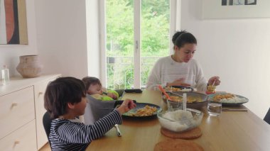 Anne yemek masasında bebeği beslerken büyük çocuk yemek yiyor, aile yemeği yiyor, ev içinde huzurlu bir ortam sağlıyor, anne çoklu görev yapıyor, yüksek sandalyedeki bebek, aile bakımı...