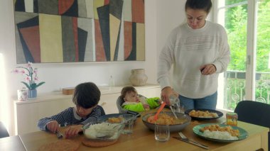 Anne yemek masasında çoklu görevdeyken, bebek yüksek sandalyede otururken ve çocuk tabağına odaklanmışken, aile yemeğine hazır.