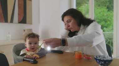 Anne yemek masasında bebeği besliyor, bebek heyecanla gülümsüyor, anne ve bebek arasındaki neşeli etkileşim, neşeli beslenme anı, aile yemeği, bebeğin yemekten zevk alması.
