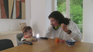 Anne yemek masasında bebeği besliyor, bebek heyecanla gülümsüyor, anne ve bebek arasındaki neşeli etkileşim, neşeli beslenme anı, aile yemeği, bebeğin yemekten zevk alması.