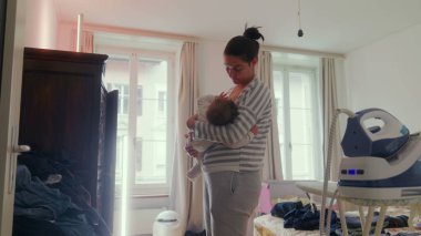 Anne bebeğini emzirirken huzurlu bir ev ortamında duruyor, sessiz beslenme anı, yakın aile bağı, anne bakımı ve evde şefkat.