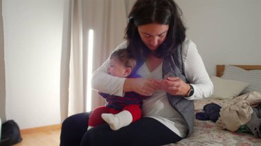 Anne yatakta oturup bebek tırnağı kesiyor, gerekli bakım ve bakımı sağlıyor, sıcak bir ev ortamında samimi ebeveynlik anları yaşıyor, bebek hijyenine odaklanıyor..