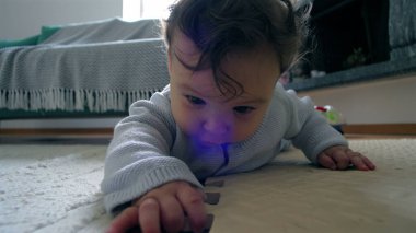 Bebek yerde yatıyor, yüzü yere yakın, tahta bir blokla etkileşimde, merakla çevreyi keşfediyor, ev ortamında güzel motor beceriler geliştiriyor.