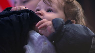 Bebek ceketi çiğniyor, yakından bakıyor, tuhaf bir ifade takınıyor, kışlık ceket giyiyor, günlük aile anı, beslenme ve koruyucu sahne, ev ortamında rahat.