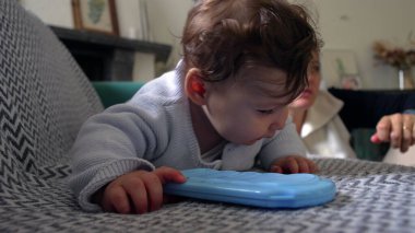 Kanepede uzanan bebek mavi nesneye uzanıyor, neşeli ve meraklı ifade, bebek oynuyor ve keşfediyor, annesiyle yakınlarda vakit geçiriyor, rahat ev ortamında eğlenceli anlar yaşıyor.