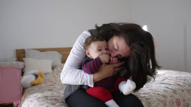 Anne neşeyle bebeği öpüyor, yatakta sevgi dolu ve neşeli anları paylaşıyor, aile evinde samimi ve eğlenceli etkileşimler kuruyor, bebek gülümsüyor ve anneyle bağ kuruyor.