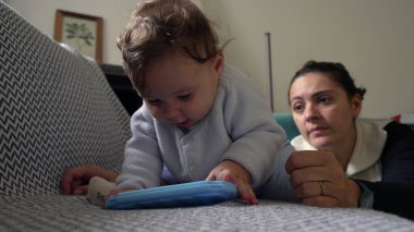 Bebek koltukta uzanmış mavi bir nesneyi tutarken, anne objeye odaklanıyor, oyun oynayarak öğreniyor, evde ailece vakit geçiriyor, anne özenle izliyor.