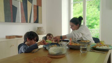 Anne yemek masasında servis yapıyor, bebek yüksek sandalyede ve çocuk yemek yiyor, aile yemeği sıcak bir evde.