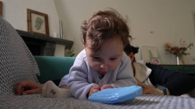 Bebek, mavi nesneyle oynarken koltukta öne eğiliyor, odaklanmış ifade, merakla bebek keşif nesnesi, kapalı alanda eğlenceli bir an, yakınlarda annesiyle aile bağı kurma zamanı.