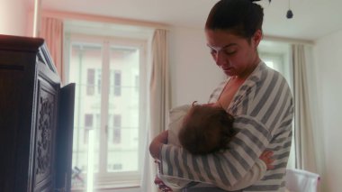 Anne, bebeğini yatak odasında durup emzirirken huzur dolu bir yuva ortamında huzur verici, şefkatli bir an yaşıyor.