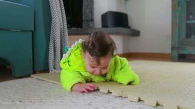 Bebek yeşil bir kıyafetle yerde yatıyor, etrafı keşfediyor, yumuşak bir paspasın üzerinde karnını inceliyor, meraklı ve meşgul, kapalı bir oyun zamanı.