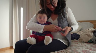 Anne bebek tırnaklarını keserken yatakta oturuyor, bebek hijyenine bakıyor, evde birden fazla iş yapıyor, gerekli özen ve ilgiyi sağlıyor..