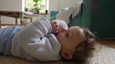 Bebek yerde birlikte yatıyor, meraklı bir ifadeyle yukarı bakıyor, evde sakin bir aile zamanı, bebek dinleniyor ve etrafı gözlemliyor, evde oyuncu bir çocuk var.