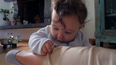 Bebek, anne bacağının üzerinde tuhaf bir ifadeyle dinleniyor, evde aile bağlarını kurma zamanı, bebek ve anne arasında oyuncu ve besleyici bir an, dokunsal bir merakla dünyayı keşfediyor..