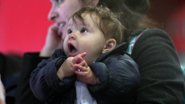 Annenin kollarında bir bebek, ceketli, heyecanlı bir yüz ifadesi, samimi bir ilişki, günlük aile anı, çevreyi gözlemleme, yakın görüş.