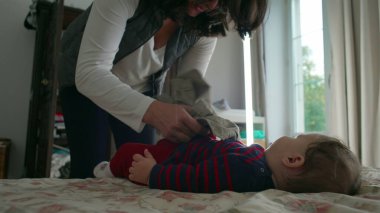 Anne bebek elbiselerini değiştirirken bebek yatakta yatar, hassas ve dikkatli bir şekilde bakım yapar, ev ortamında bebek hijyenine ve hijyene dikkat eder..