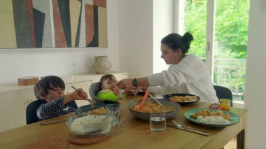 Anne yemek masasında servis yapıyor, bebek yüksek sandalyede ve çocuk yemek yiyor, aile yemeği sıcak bir evde.