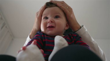 Anne bebeğin başını nazikçe tutuyor, bebek kucağında otururken neşeli bir etkileşim, aile evinde bir bağ kurma anı, ebeveynler ve çocuklar arasında bir bağ..