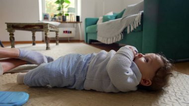 Bebek ağzında parmaklarıyla sırt üstü uzanıyor, sakin ve rahat bir yüz ifadesi, evde aile zamanı, bebek gezisi çevresi, huzurlu bir an, rahat bir evde neşeli bir çocuk.