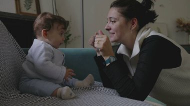 Anne bebekle kanepede iletişim kuruyor, şakacı yüz yüze iletişim kuruyor, erken çocukluk gelişiminde neşe ve bağlılık gösteriyor, ev ortamını büyütüyor.