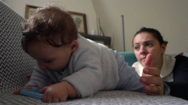 Bebek koltukta uzanmış mavi bir nesneyi tutarken, anne objeye odaklanıyor, oyun oynayarak öğreniyor, evde ailece vakit geçiriyor, anne özenle izliyor.