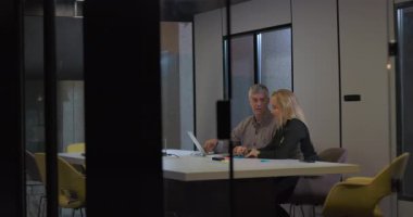 İki iş adamı, cam duvarlı konferans masasında akşam görüşmelerini paylaşıyor, notlar ve dizüstü bilgisayarları incelerken iş görüşmesi yapıyorlar.