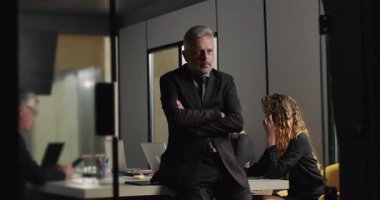 Üst düzey işadamı, ofis yönetim kurulunda düşünceli bir duruş sergilerken, iş arkadaşları şirket toplantısında dizüstü bilgisayarlara yoğunlaşıyor.