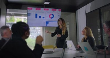 Meslektaşlarına veri tablosu sunan genç bir kadın. Bu sırada sarışın iş kadını ve ekip üyeleri, iş birliği bürosu toplantısını dikkatle dinliyorlar.