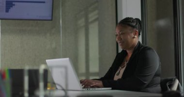 Modern ofis ortamında sessiz çalışma saatlerinde dizüstü bilgisayarla yazarken hafifçe gülümseyen Afrikalı Amerikalı iş kadını.