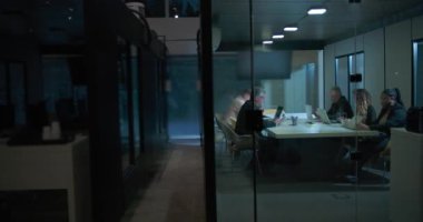 Şirketin gece vardiyası sırasında cam toplantı odasında laptoplar ve belgeler masaya serilerek geç saatlere kadar çalışan çeşitli takımların geniş görüşü.